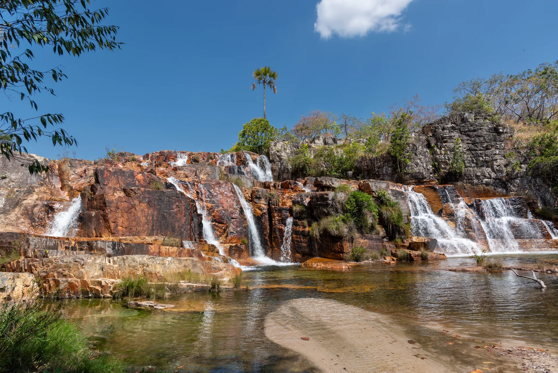 Quedas d'água em degraus de pedra avermelhada, com uma palmeira no alto e um banco de areia no poço, na Cachoeira Boa Brisa