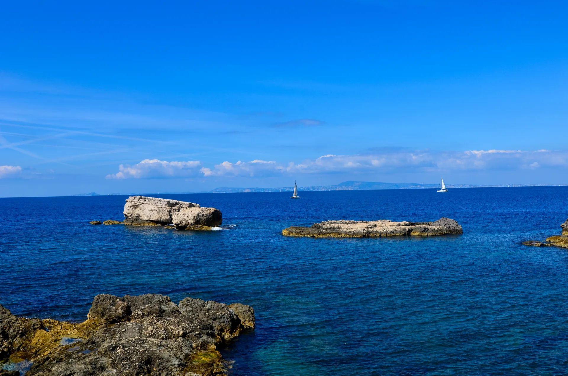 Rochedos claros no mar azul do Mediterrâneo, com dois veleiros no horizonte, em Capo di Sorrento