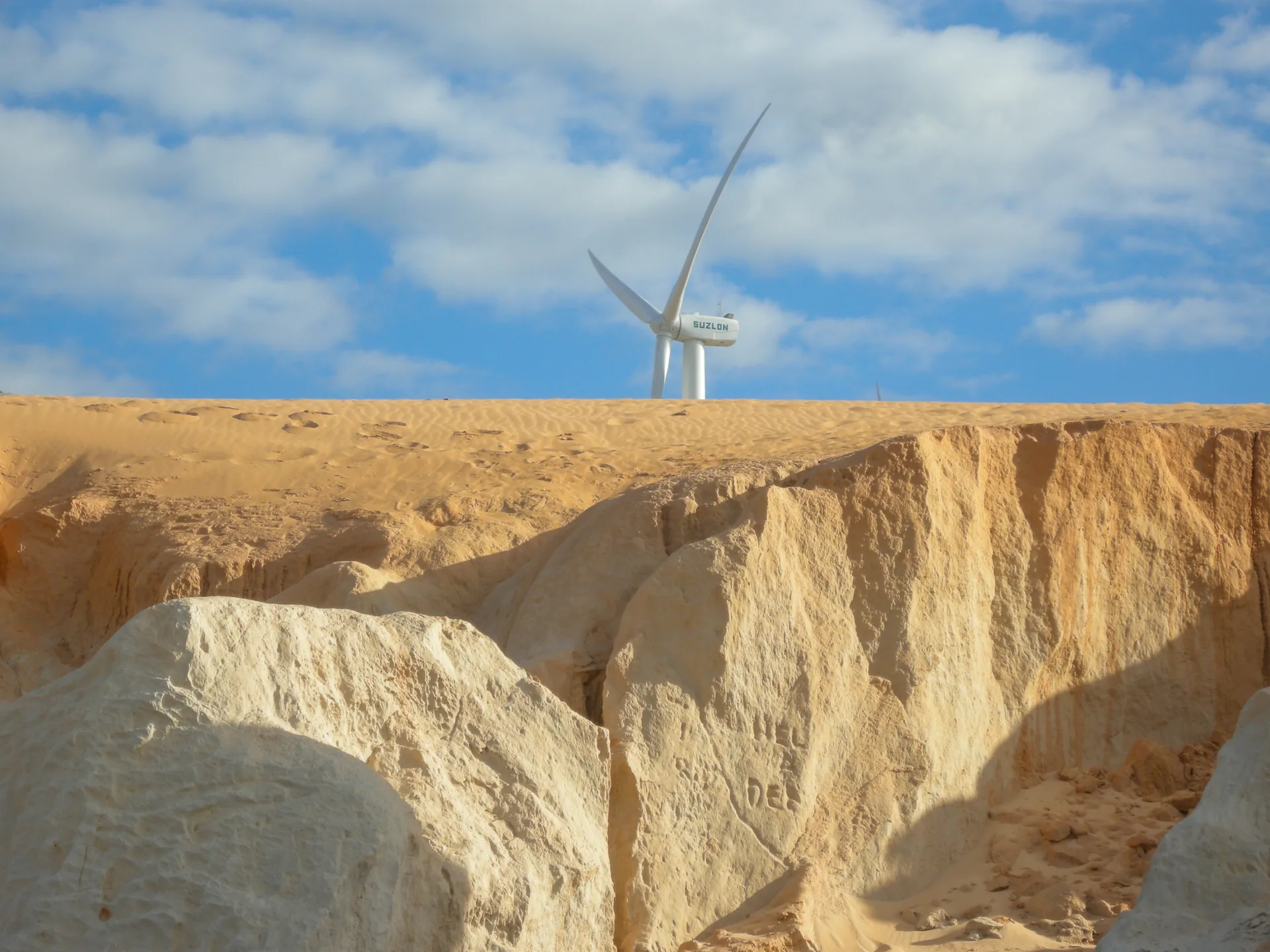 Torre de energia eólica atrás de falésias de areia clara, sob céu com nuvens, em Canoa Quebrada