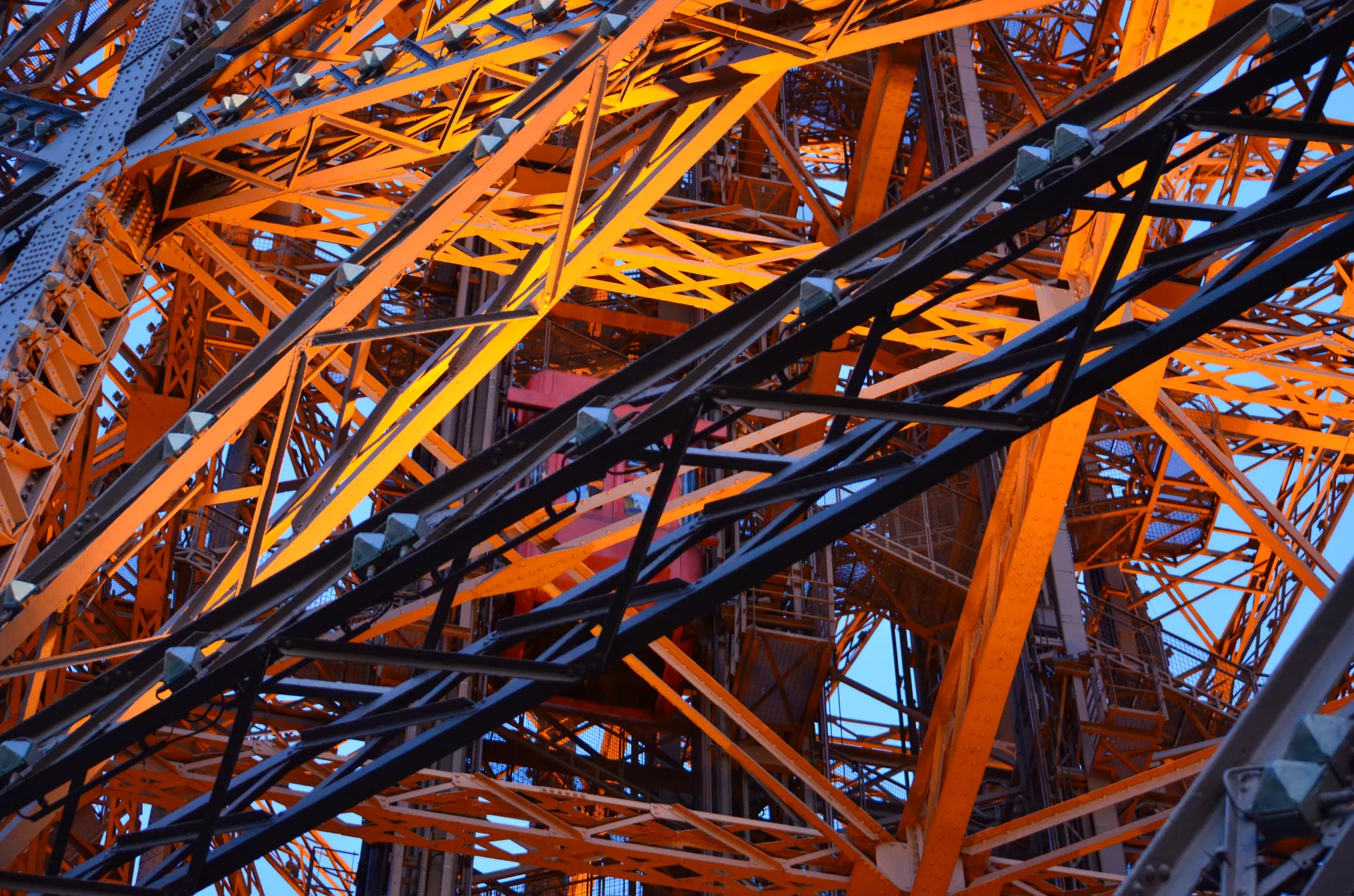 Vigas de ferro entrelaçadas da Torre Eiffel, iluminadas em tons de laranja, vistas de baixo