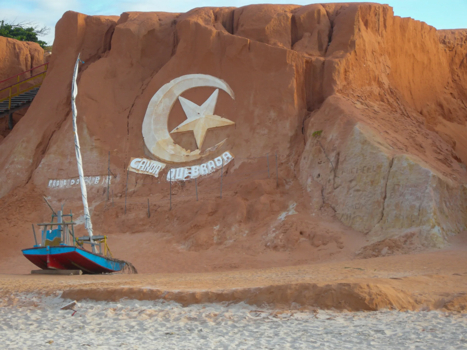Falésia avermelhada com o símbolo da lua e da estrela e uma jangada na areia, em Canoa Quebrada