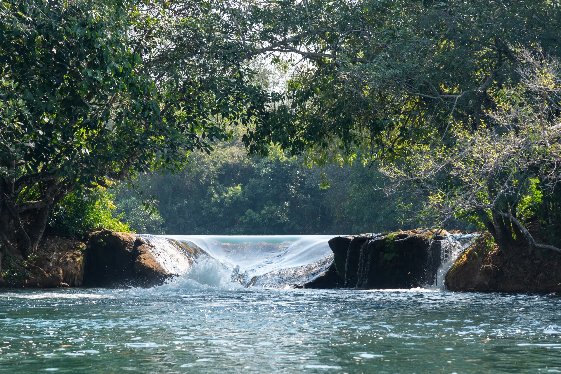 Pequena cachoeira entre pedras escuras, com água verde-azulada e árvores em volta, no Rio Formoso, em Bonito
