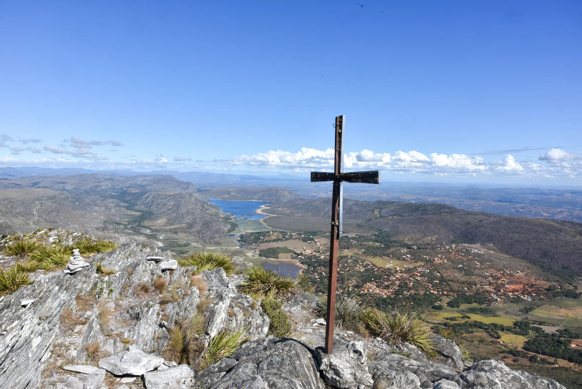 Cruzeiro de madeira no alto de rochas claras, com vale, represa e serras ao fundo, no Pico da Lapinha