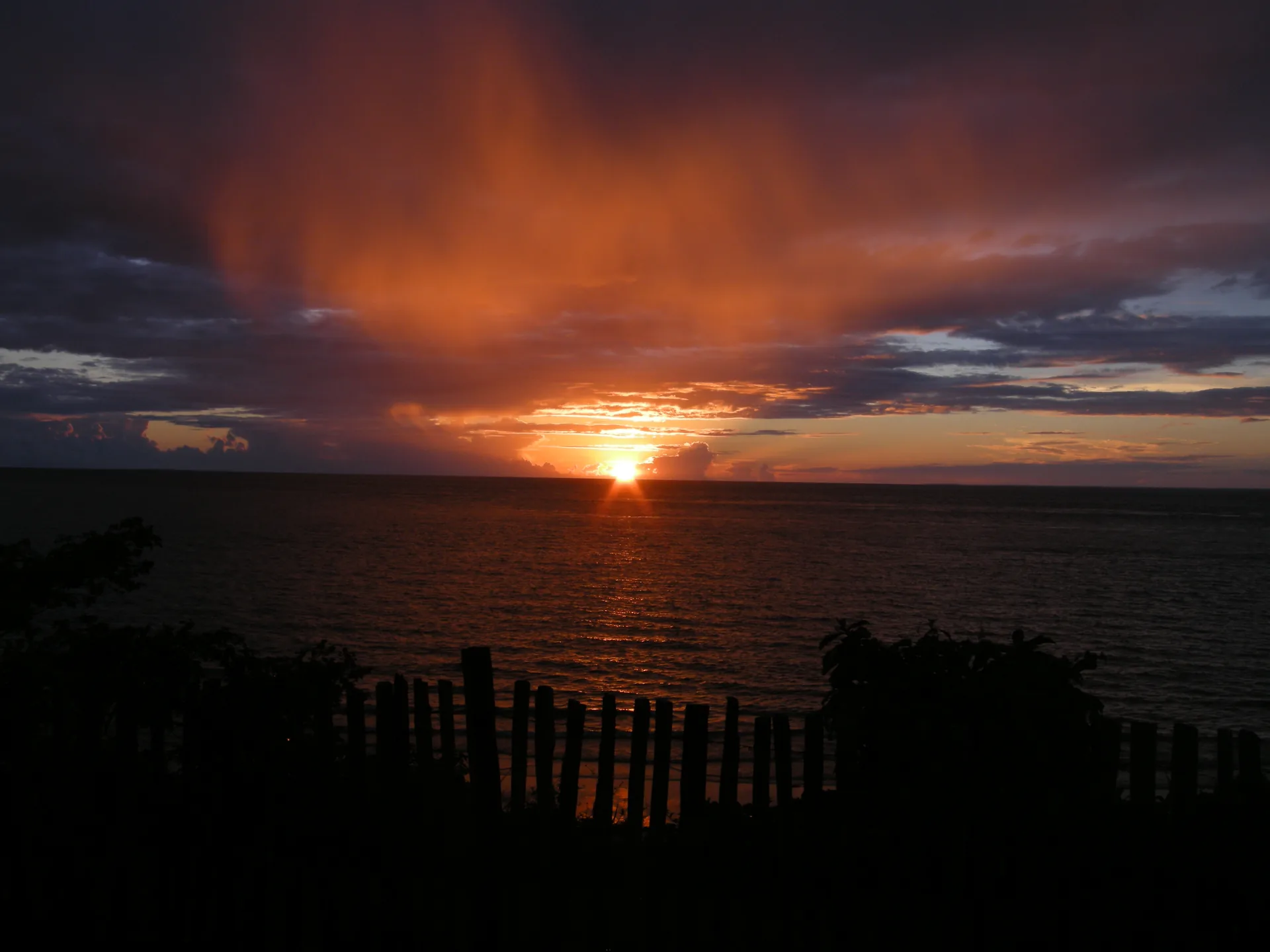 Sol se pondo no horizonte da água sob nuvens alaranjadas, com a silhueta de uma cerca em primeiro plano, em Soure