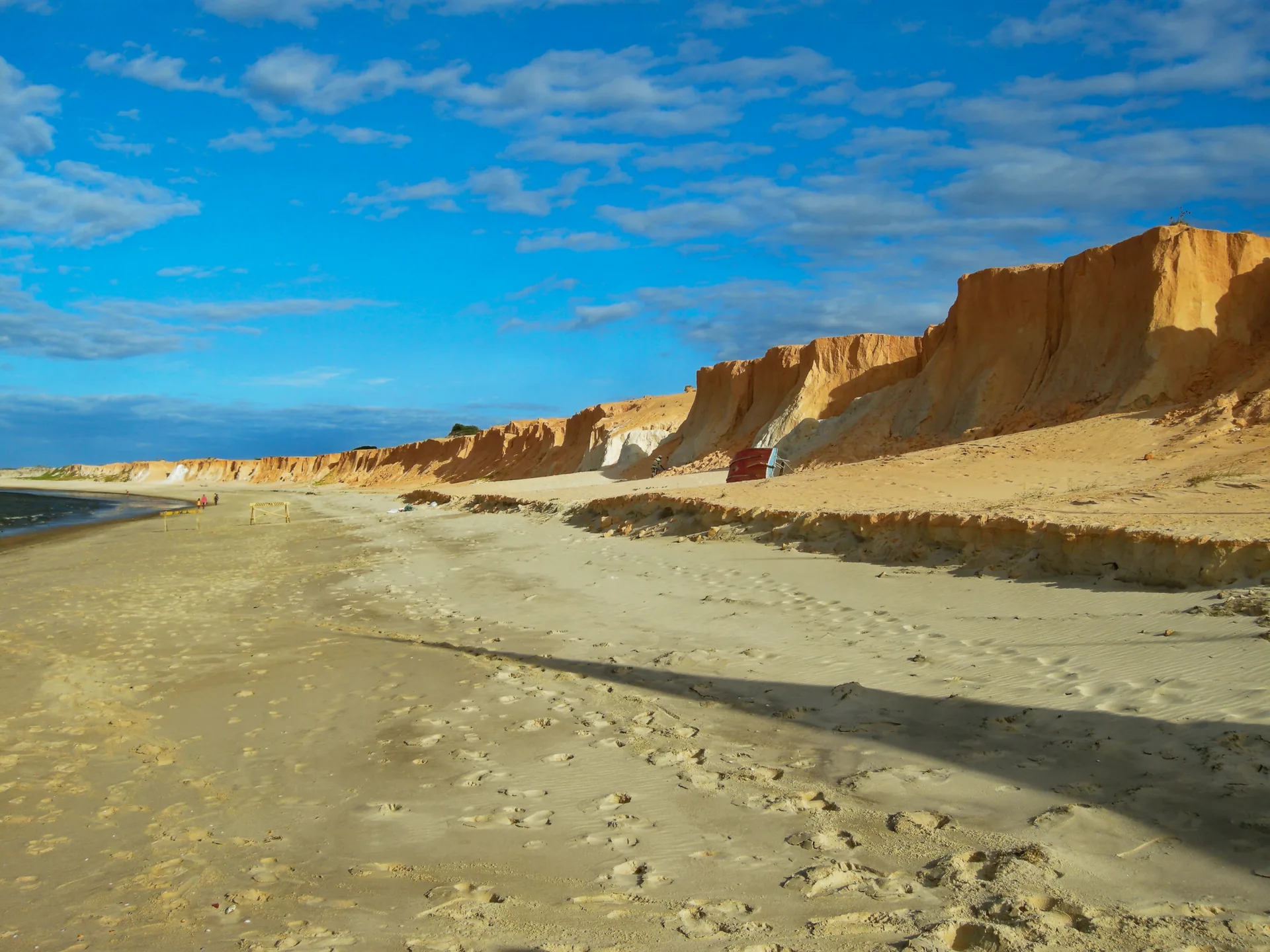 Faixa de areia clara ao pé de falésias avermelhadas, sob céu azul, na praia de Canoa Quebrada