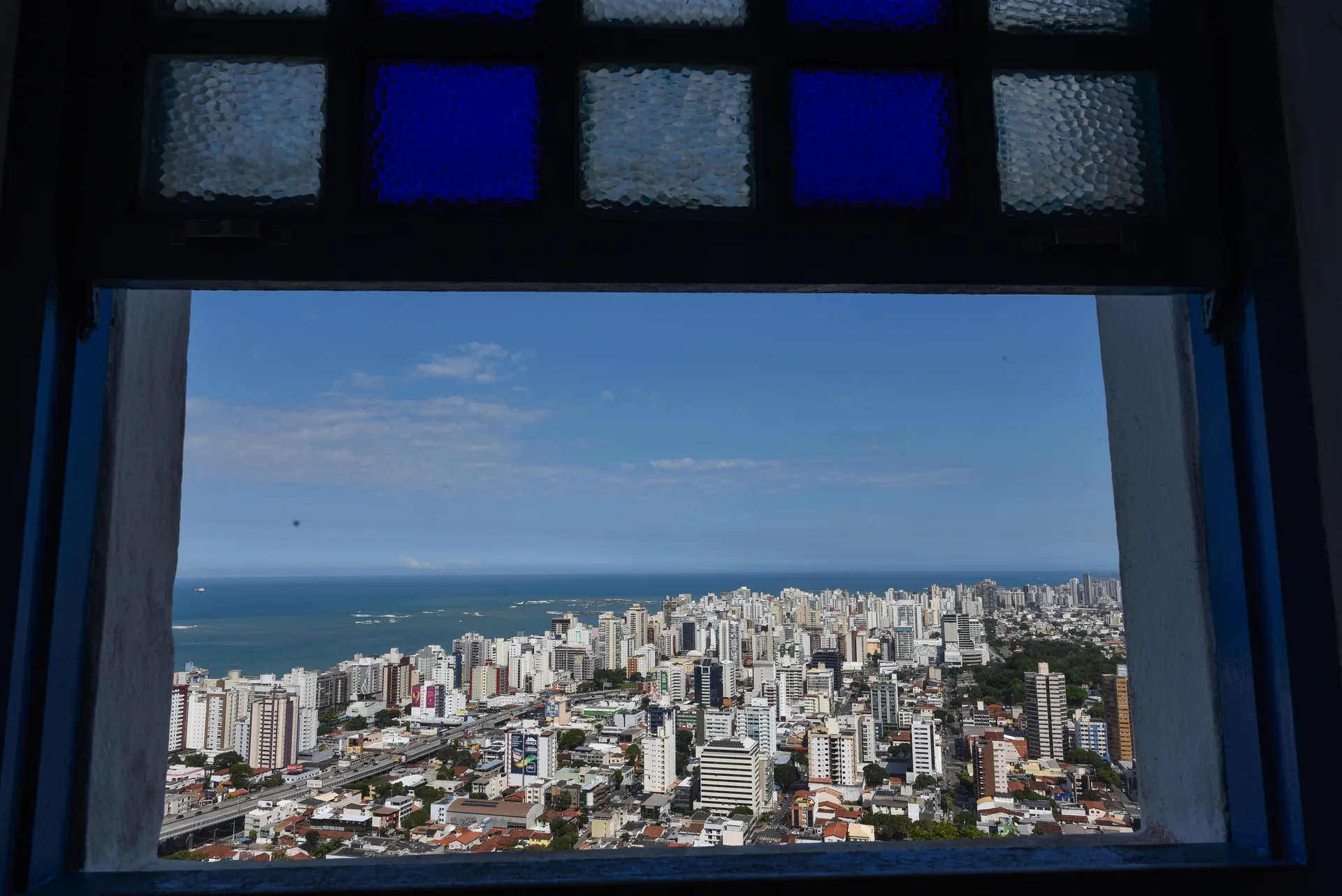 Prédios de Vila Velha e o mar vistos pela janela do Convento da Penha, com vitrais azuis no alto